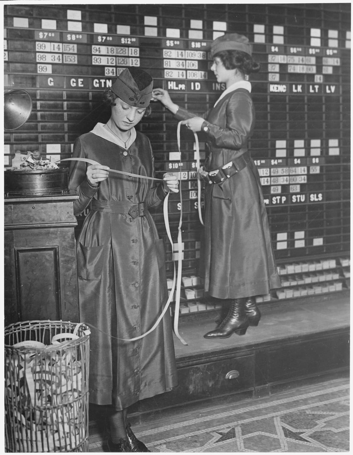 Two women in uniform in front of a wall-sized stock board: one reads a strip of ticker tape, the other stands on a step posting prices under the tickers.