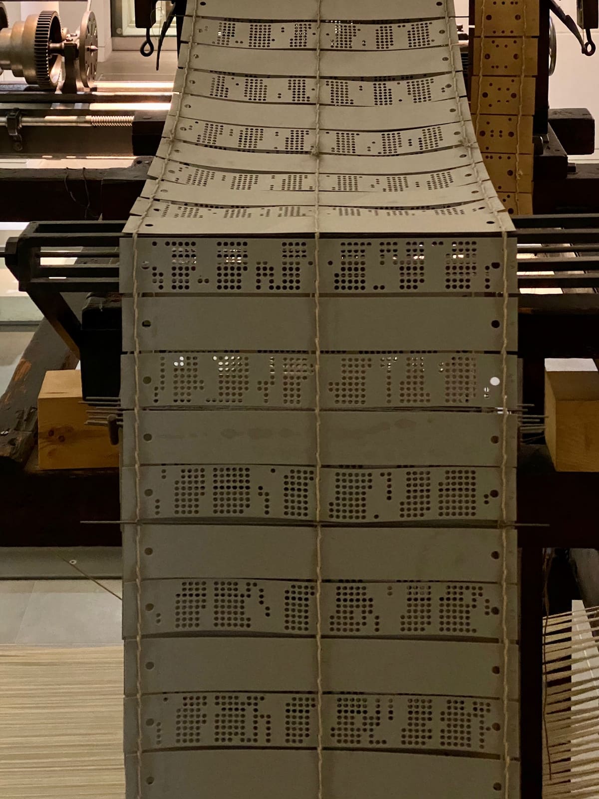 A long chain of punched cards laced together with string, hanging from the top of a Jacquard loom in a museum.