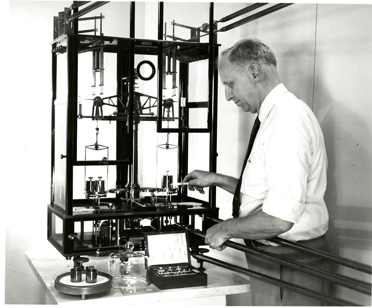 A man at a glass-cased precision balance at the U.S. National Bureau of Standards, a box of reference weights beside it.