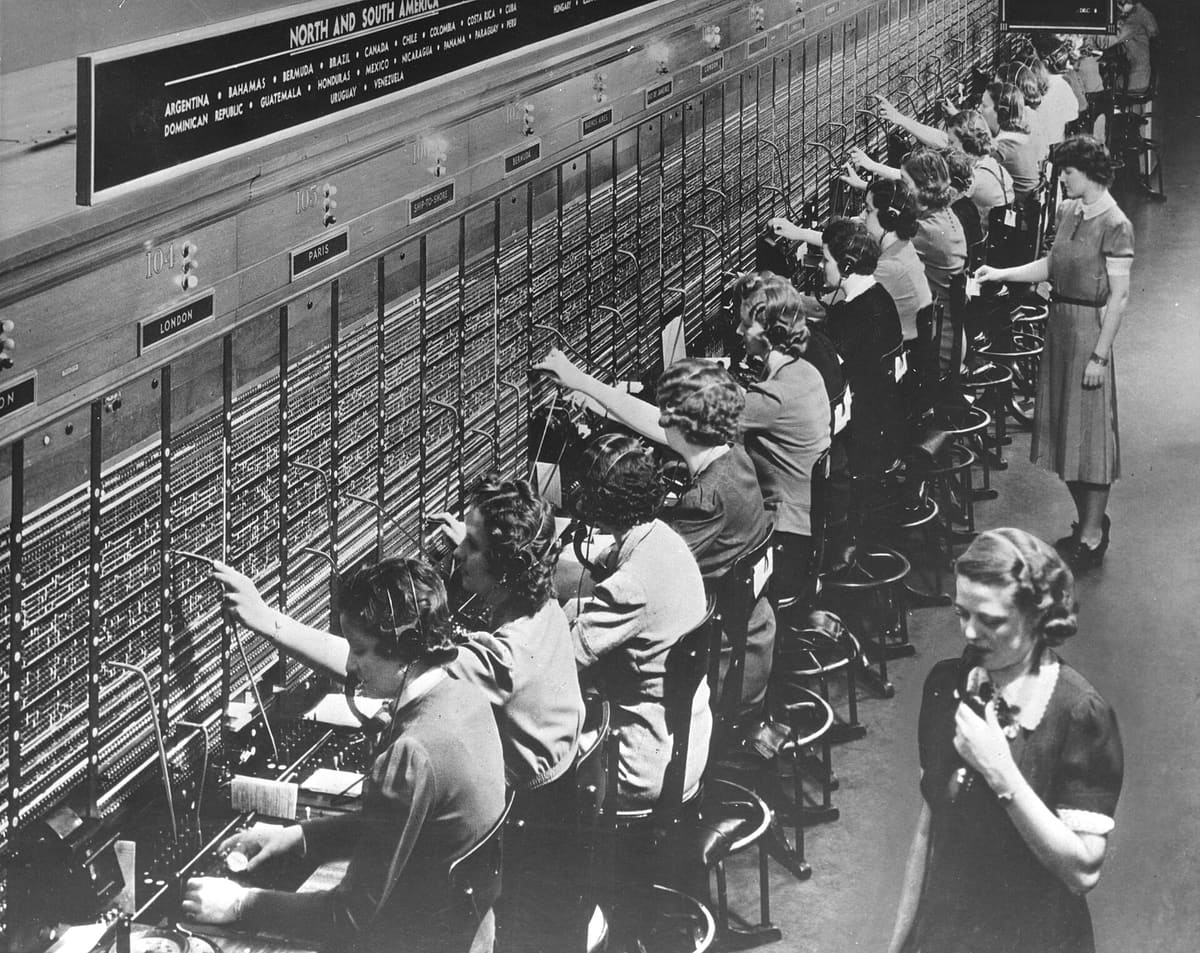 A long row of operators in headsets at a Bell System international telephone switchboard, its sections labelled London, Paris, Bermuda and Ship-to-shore.