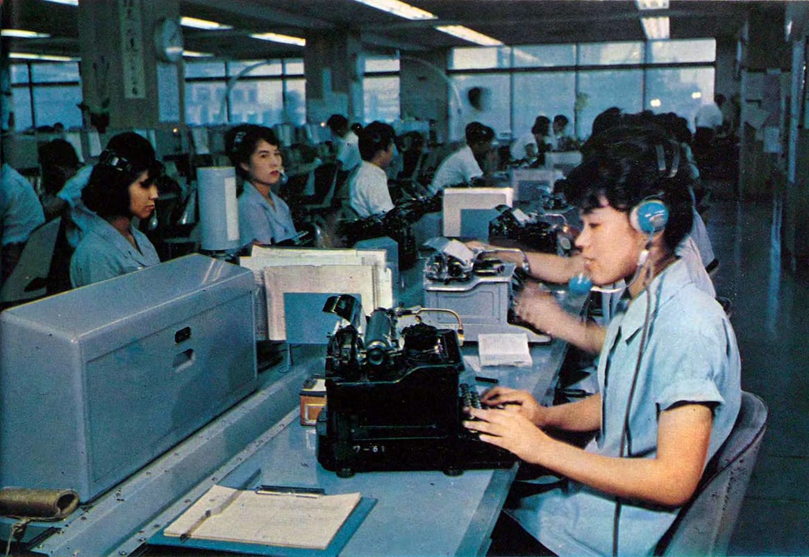 Operators in headsets typing incoming telegrams on teleprinters along a long desk at the Tokyo Central Telegraph Office, 1963.